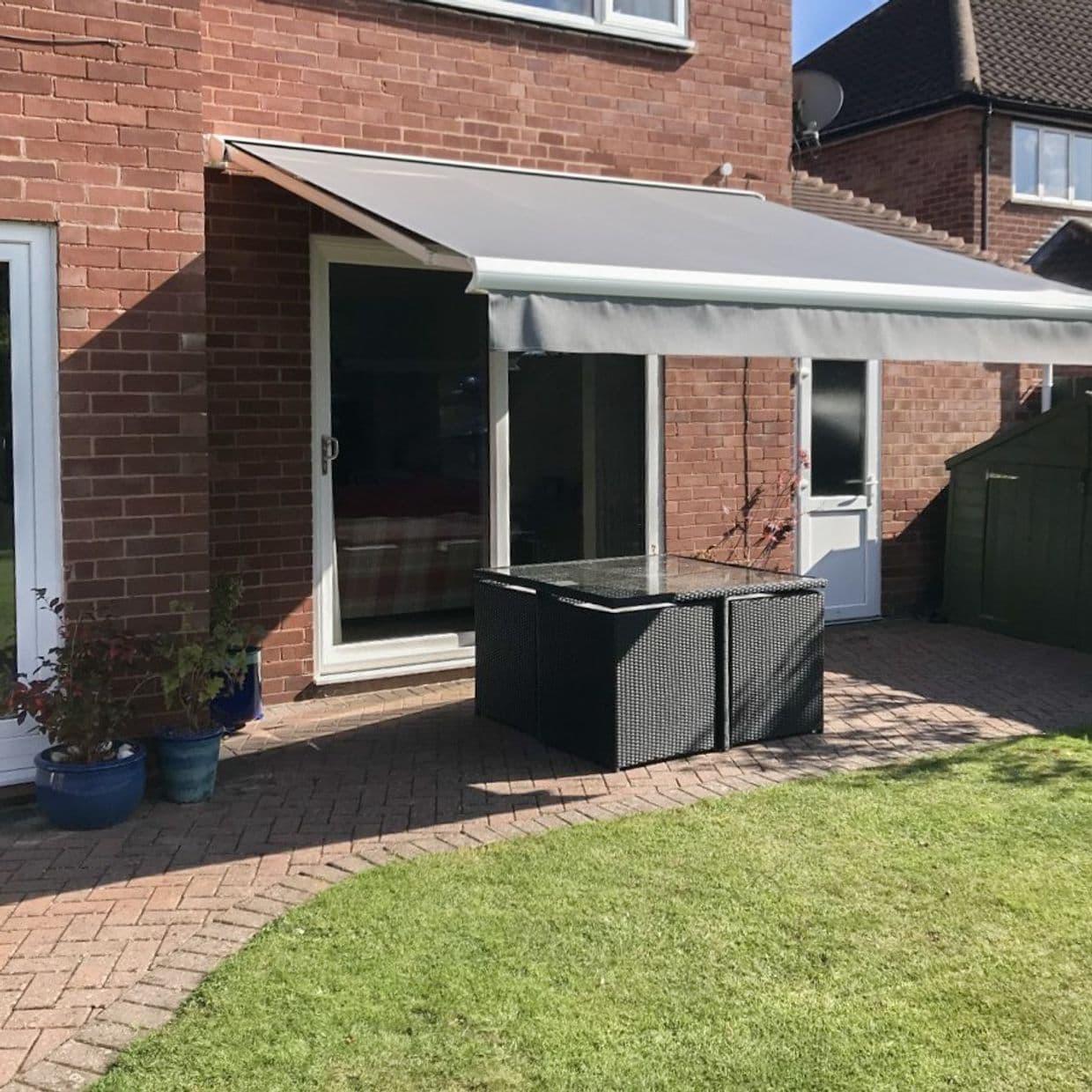 A grey retractable awning extended over a sliding glass door, shading a patio; rattan table beneath on brick pavers outside a red-brick house, sunny lawn in foreground.