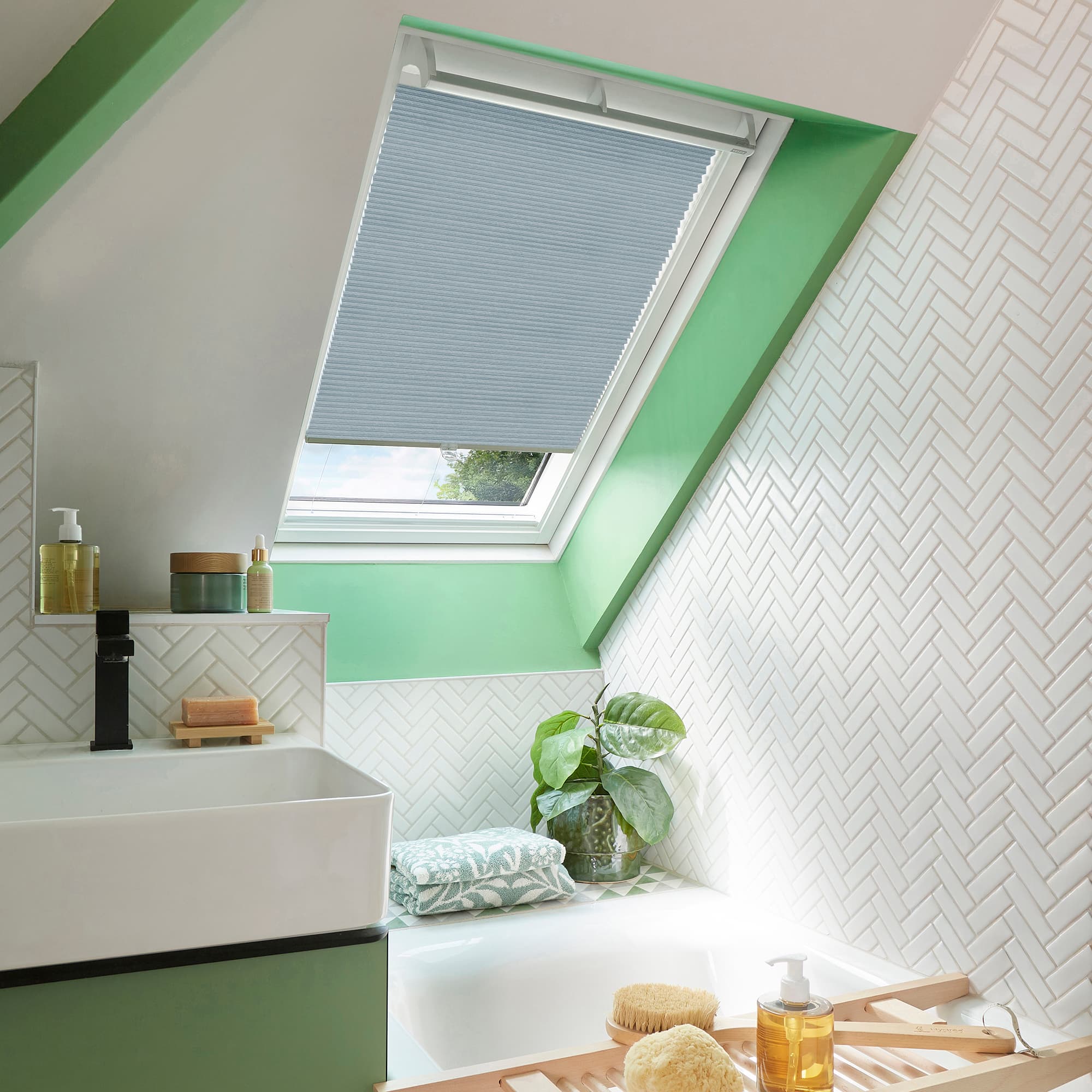 A light-grey pleated skylight blind, partially lowered and filtering daylight, covers a roof window; inside a bright attic bathroom with green-painted trim, white herringbone tiles, sink, bathtub, and potted plant.