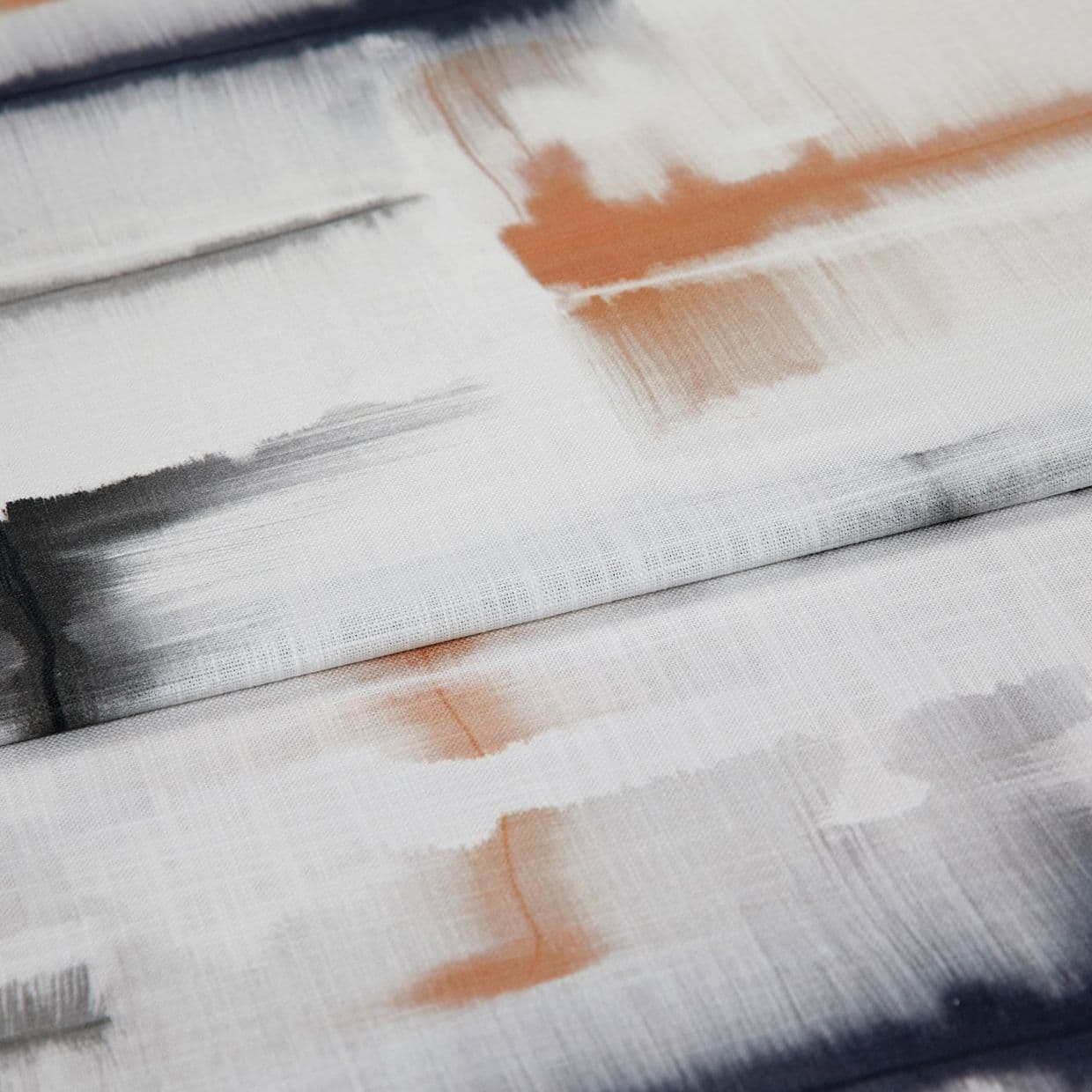Folded white fabric featuring soft watercolor-like brushstroke prints in black and rust; layered and slightly creased, showing woven texture under diffuse studio lighting in a close-up textile sample.