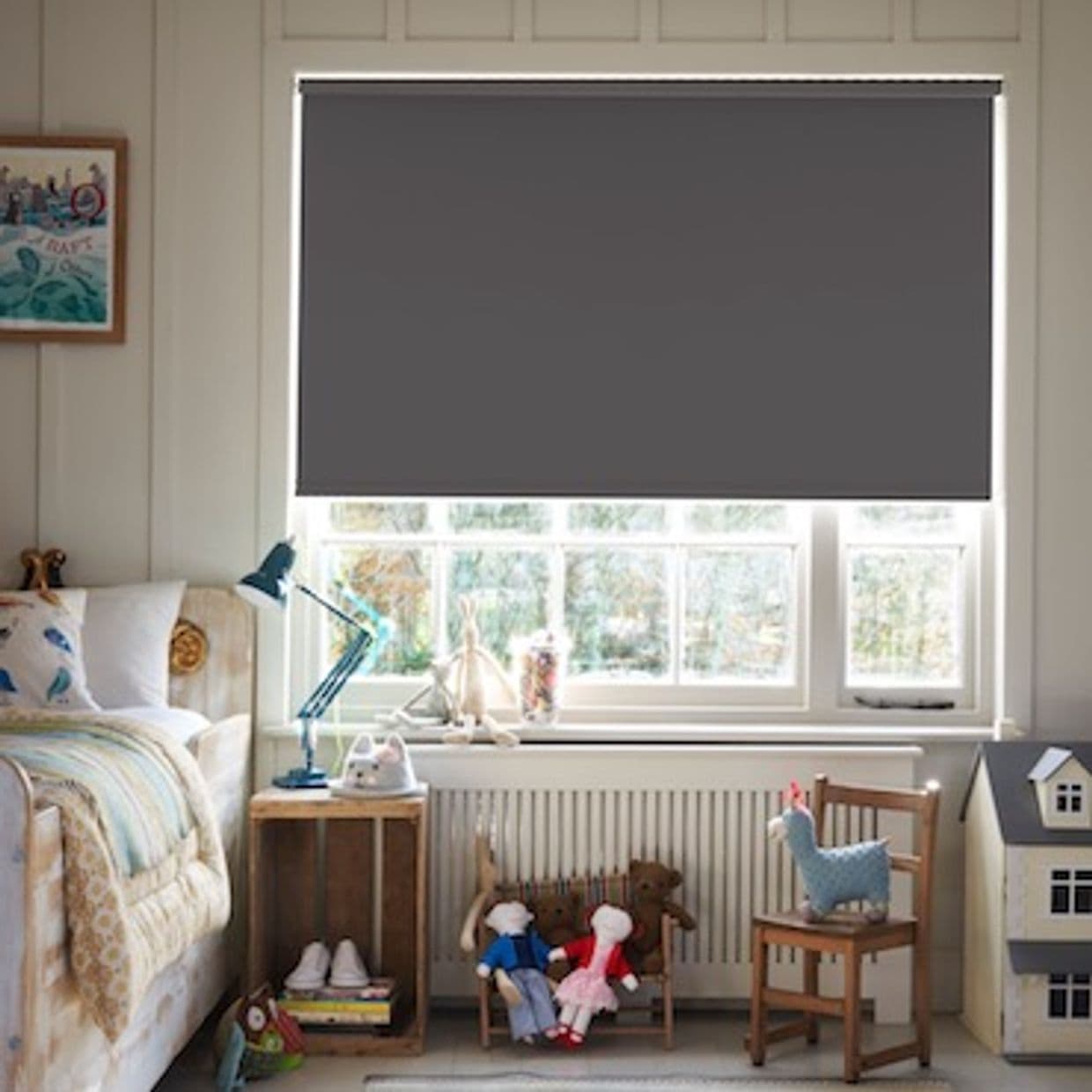 Dark gray roller blind lowered across window, edges slightly backlit. Action: blocking daylight. Context: cozy children's bedroom â€” bed, bedside table and lamp, dolls and toy house on chairs beneath window.