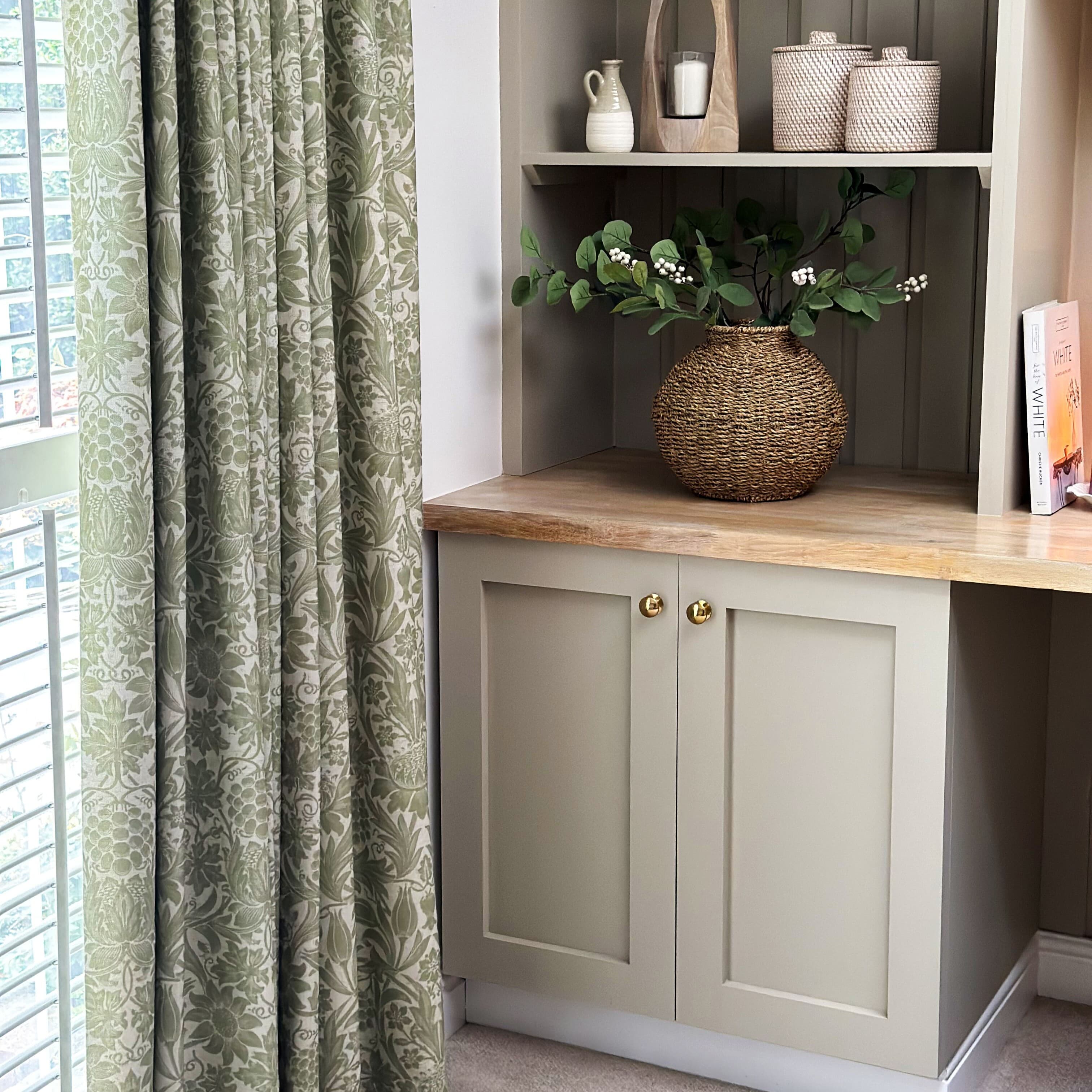 Green patterned pleated curtains at left, drawn closed and filtering daylight through white blinds; beside a beige built in cabinet with wooden countertop and woven vase of greenery.