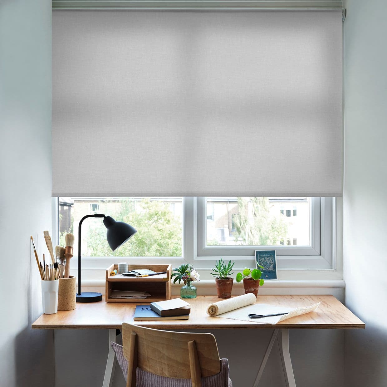 Light-gray fabric roller blind lowered across the window, diffusing daylight and partially obscuring the view; a wooden desk below holds a lamp, art brushes, plants and notebooks in an office.