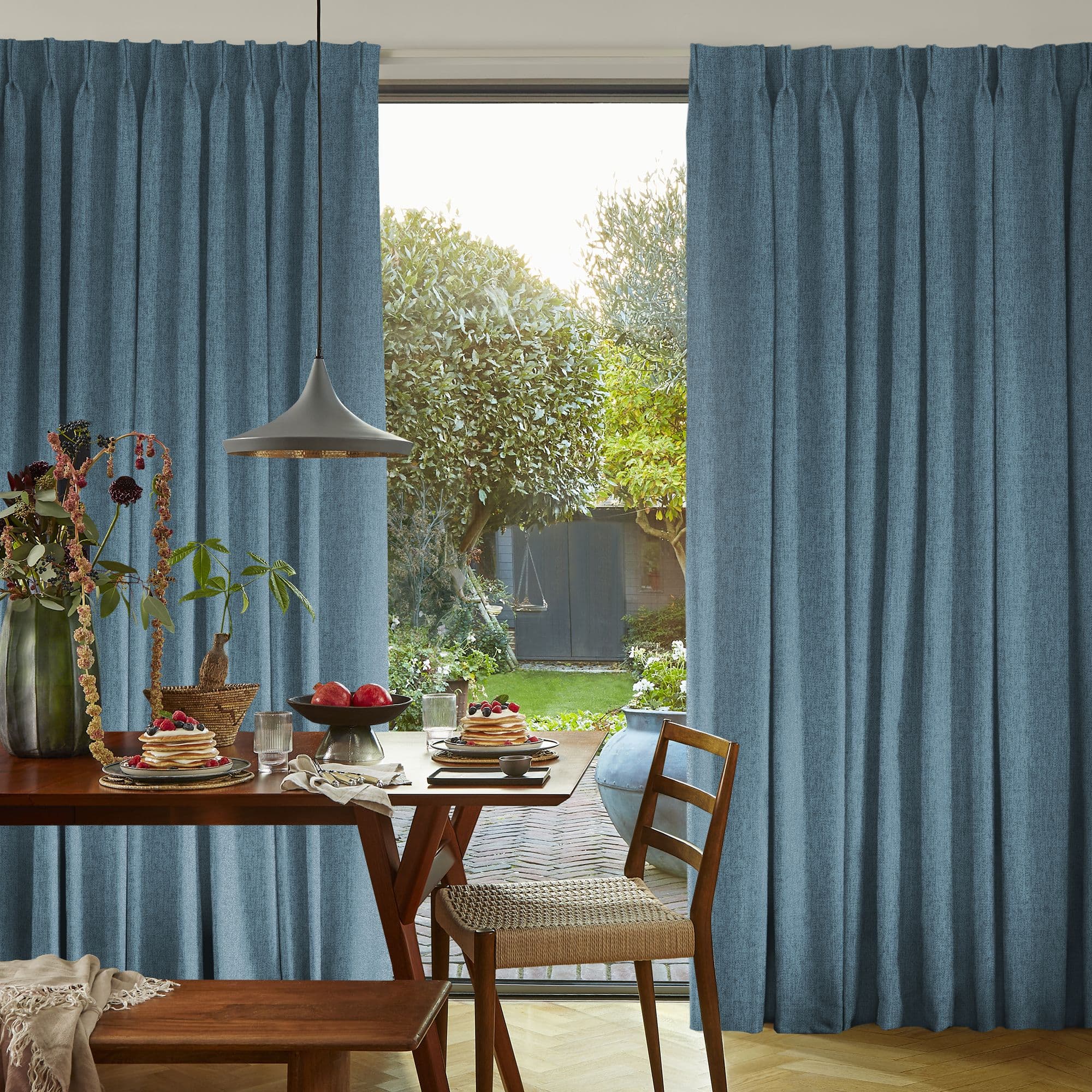 Blue pleated curtains, parted at center to admit daylight through sliding glass doors; dining table set for breakfast with wooden chairs, pendant light, and a sunlit garden visible beyond.