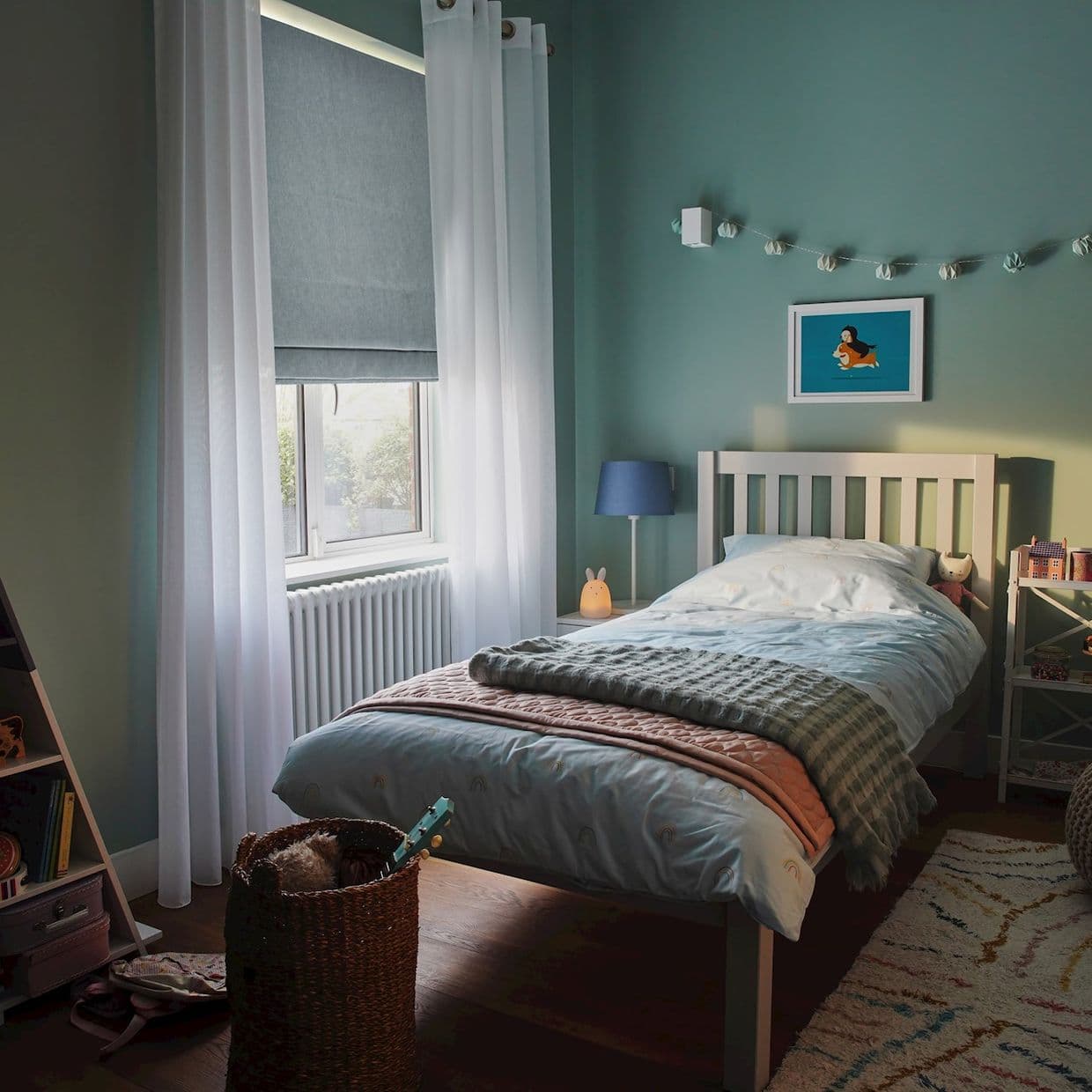 Sheer white grommet curtains flank a lowered grey Roman blind over the window, partially filtering daylight; small teal bedroom with a single bed, bedside lamp, soft toys, and rug.