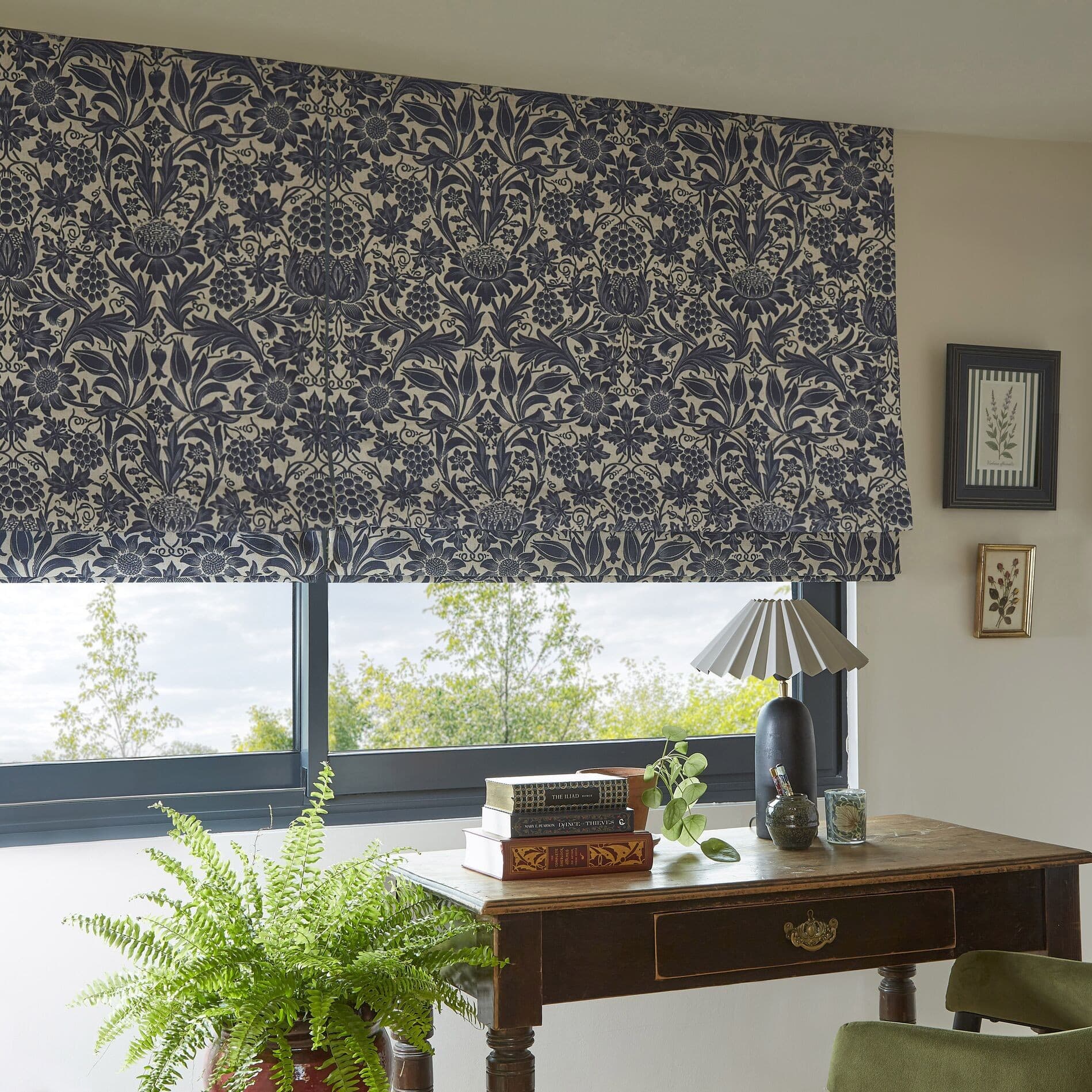 Patterned fabric Roman blind, lowered across the window with dense navy botanical print on cream, filtering daylight over a wooden desk with lamp, books, fern and a treed outside view.