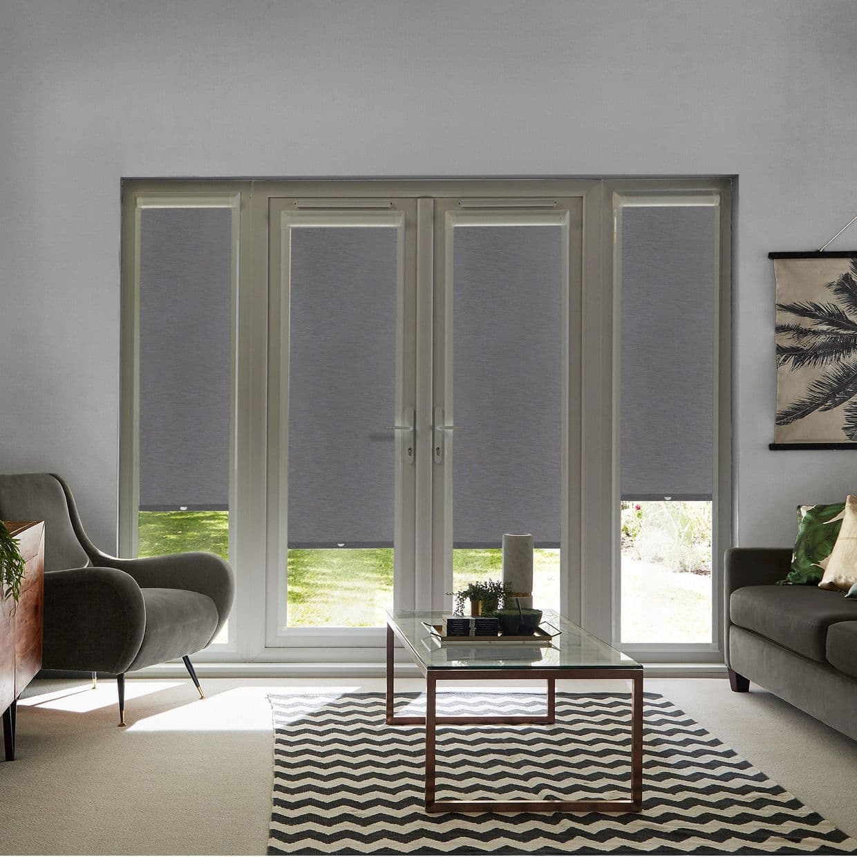 Grey roller blinds covering four glass panels of French doors and sidelights, lowered mid-height filtering daylight; living room with armchairs, glass coffee table, patterned rug and garden view.