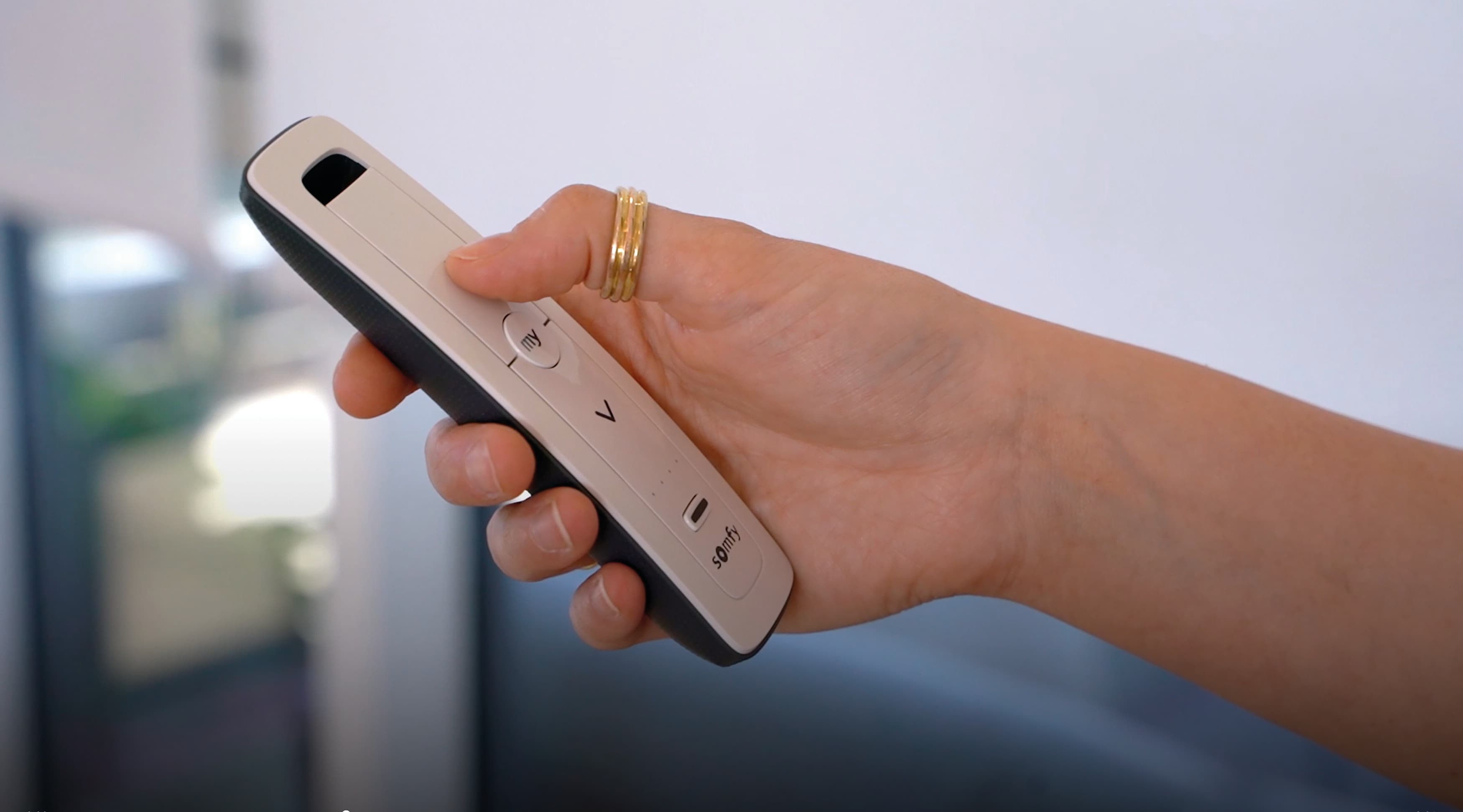 Slim white-and-black remote control held in a right hand, thumb pressing a circular my button; indoor, softly lit room with a blurred window or glass door in the background.

Text: my V somfy