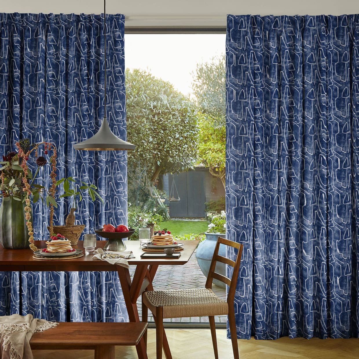 Blue pleated fabric curtains with white abstract pattern, partially drawn aside, framing a sunlit dining table and open glass door leading to a lush backyard garden.