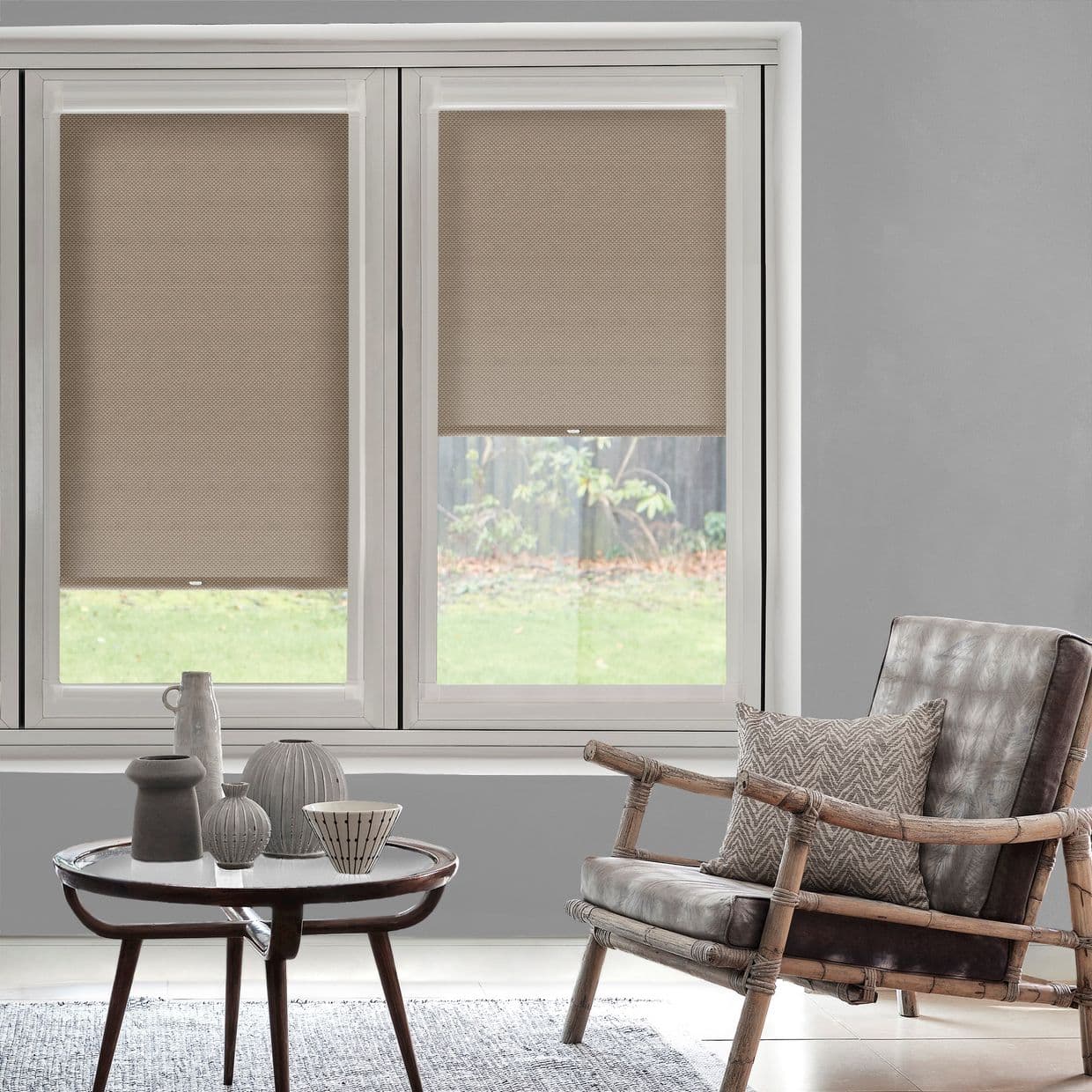 Beige textured roller blinds, partially lowered across twin-pane window, filtering daylight; softly lit living room with low round wooden table holding ceramic vases, rattan-frame leather armchair and grassy yard outside.