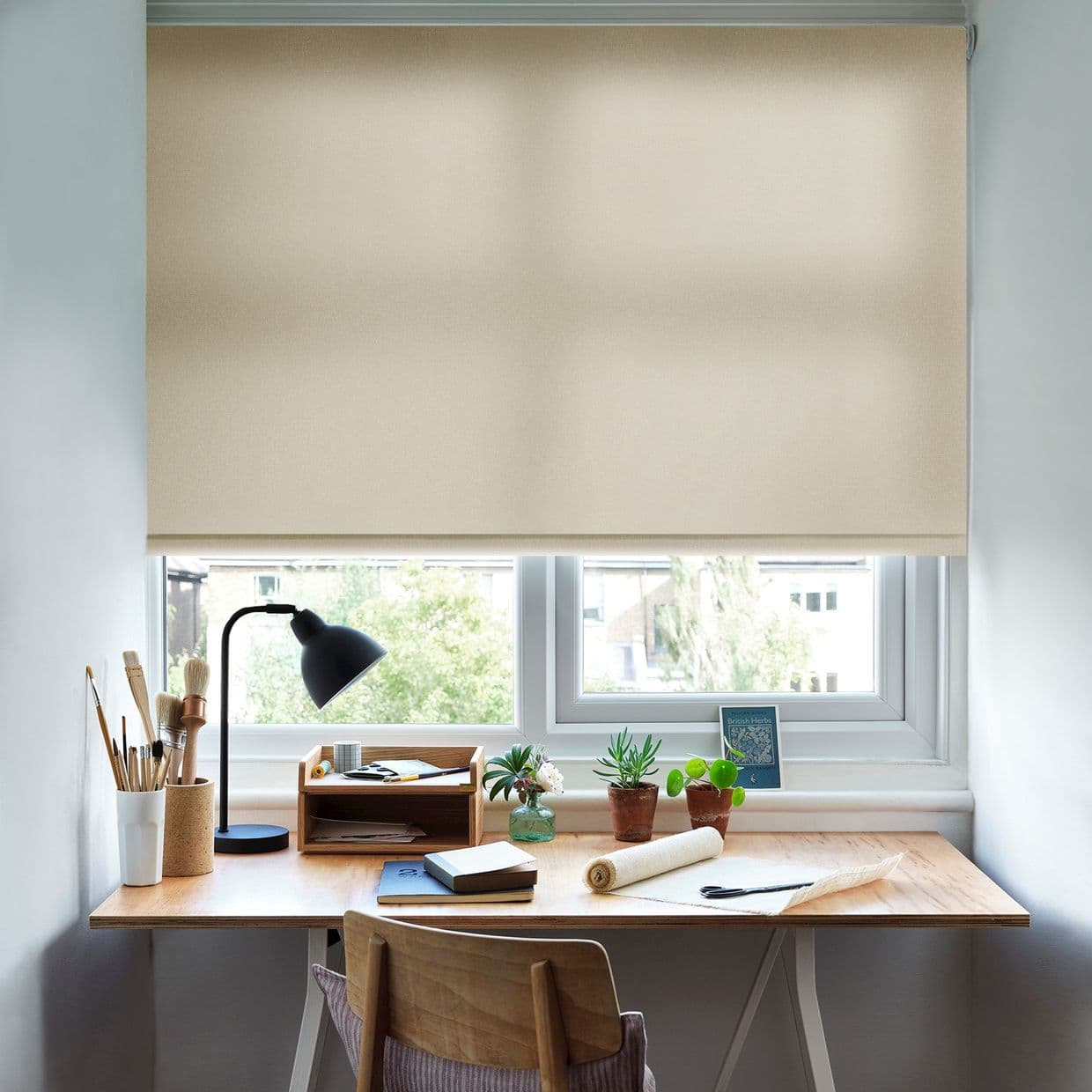 A beige fabric roller blind lowered to mid-window, slightly translucent, filtering soft daylight into a tidy wooden home workspace with desk lamp, potted plants, art brushes and notebooks, tree view.