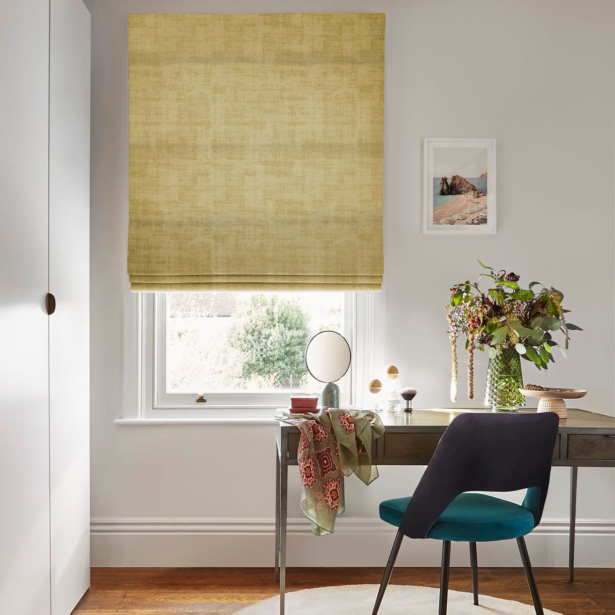 Yellow textured Roman blind lowered over window with soft folds; filtering daylight; home office desk beneath holds a round mirror, vase of flowers and scarf, teal chair, framed seaside print.