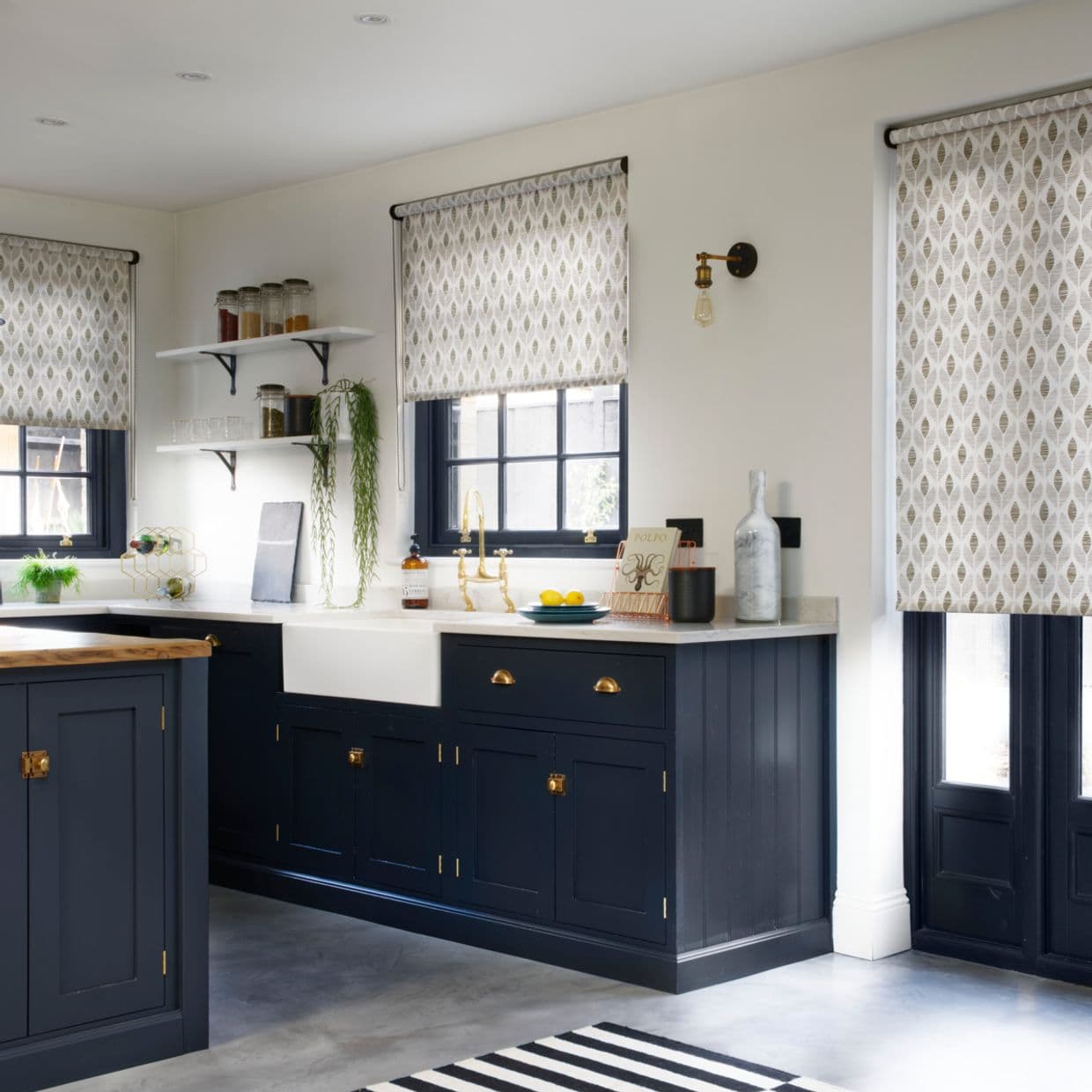 Leaf-patterned roller blinds, mounted above three windows and partially lowered, filtering soft daylight into a bright kitchen with navy lower cabinets, white countertops, farmhouse sink and brass fixtures.