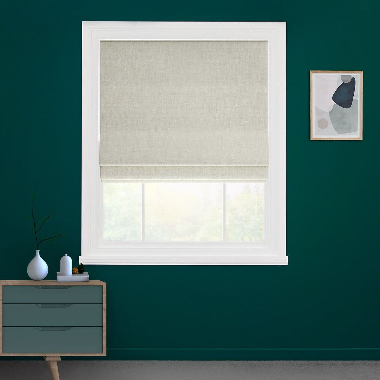 Beige fabric Roman blind, lowered with a small bottom fold across a white-framed window; filtering soft daylight; set on a teal-painted wall above a mid-century dresser and framed abstract art.