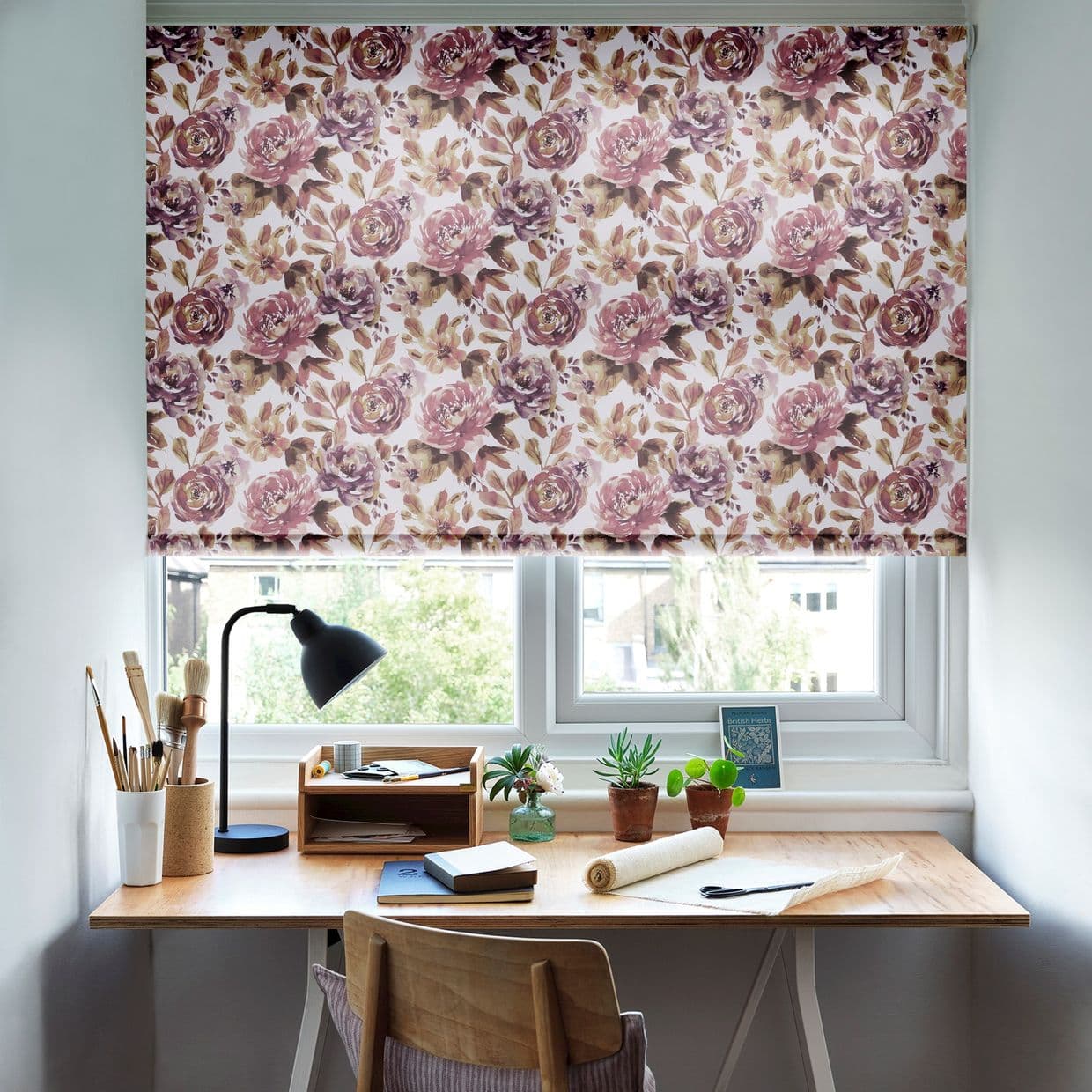 Floral fabric roller blind, lowered and filtering daylight through twin casement windows; overlooks a leafy view. Below, a wooden desk workspace with lamp, art brushes, notebooks, and potted succulents.