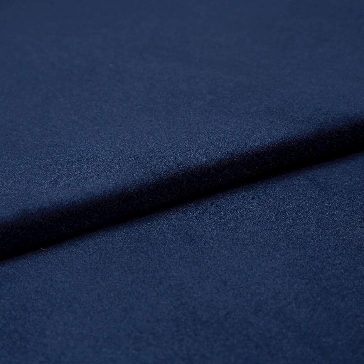 Navy-blue soft fabric, folded into overlapping layers creating a diagonal ridge and subtle shadow, showing a fine, slightly nappered texture; close-up studio view with soft, even lighting.