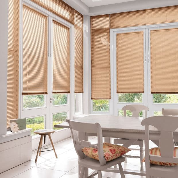 Honey colored horizontal Venetian blinds on tall bay windows, lowered and filtering daylight; white dining table and chairs in a sunlit breakfast room, small stool and green garden beyond.
