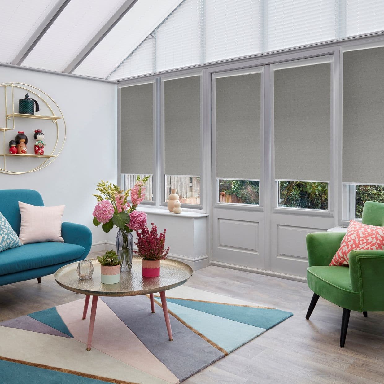 Grey roller blinds lowered over tall glazed doors and windows, textured and leaving small bottom gaps, filtering light in a bright conservatory living room with sofa and coffee table.