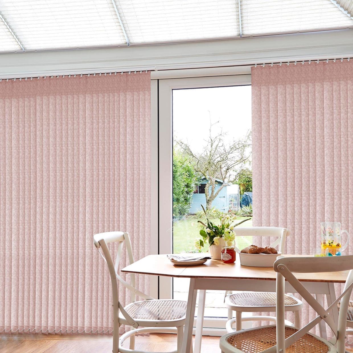Vertical pale-pink fabric blinds, floor-to-ceiling pleated panels across a sliding glass door; mostly closed, parted centrally to admit diffuse daylight; small breakfast/dining area with table, chairs, vase, and garden view.