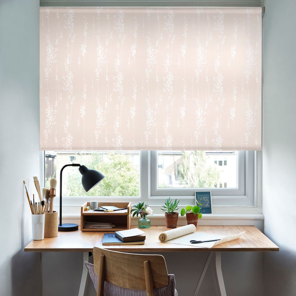 Pale pink roller blind with white vertical floral pattern, fully lowered, softly diffusing daylight, over a wooden desk with lamp, paints and potted plants in a bright home study.