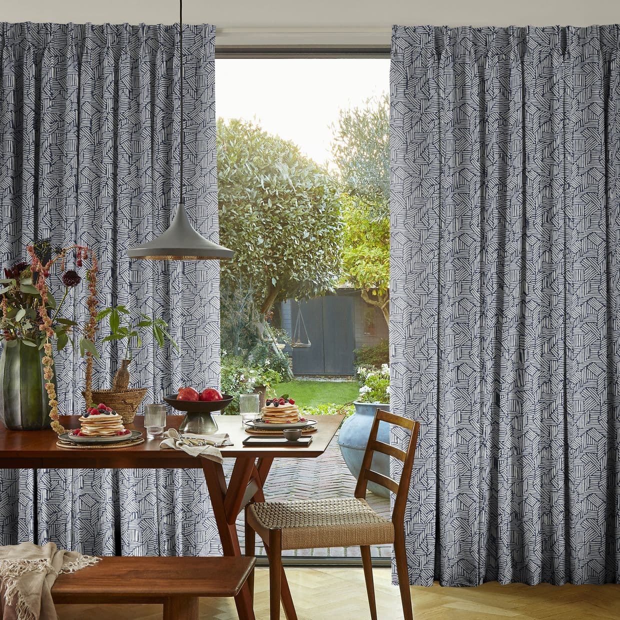 Pleated blue-and-white geometric curtains, partly drawn to reveal a sliding door; filtering daylight into a dining area with wooden table set for breakfast and a leafy garden beyond.