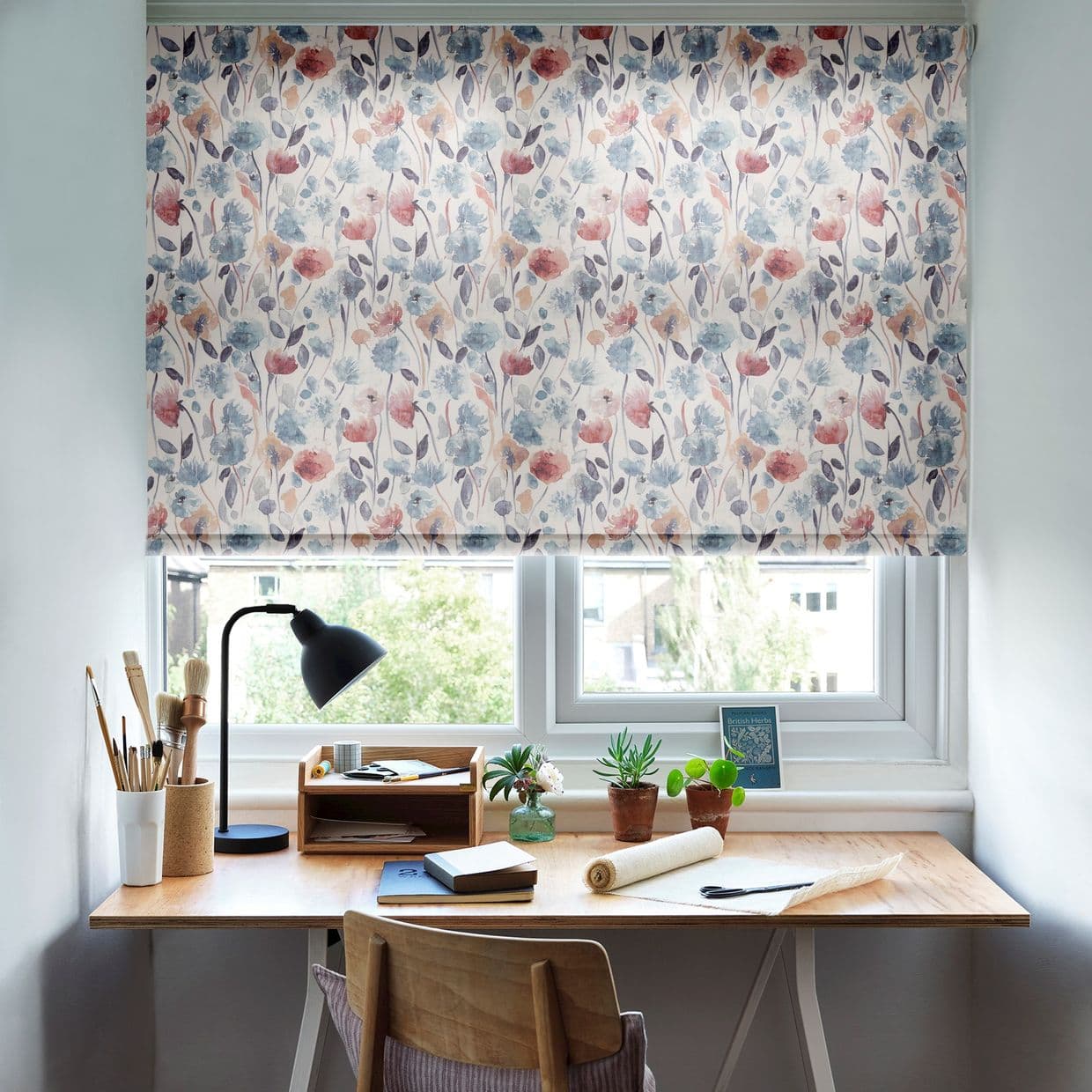 Floral roller blind lowered across the window, watercolor blueâ€‘andâ€‘pink flowers; filtering soft daylight into a small home workspace â€” wooden desk with lamp, art brushes, notebooks and potted plants.
