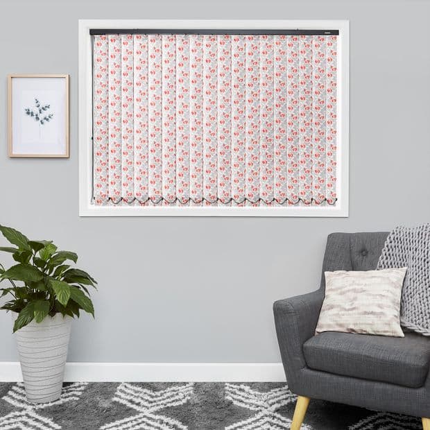 Vertical fabric blinds with pink floral print, fully closed across the window with scalloped hem; in a grey living room with potted plant, framed print, grey armchair and rug.