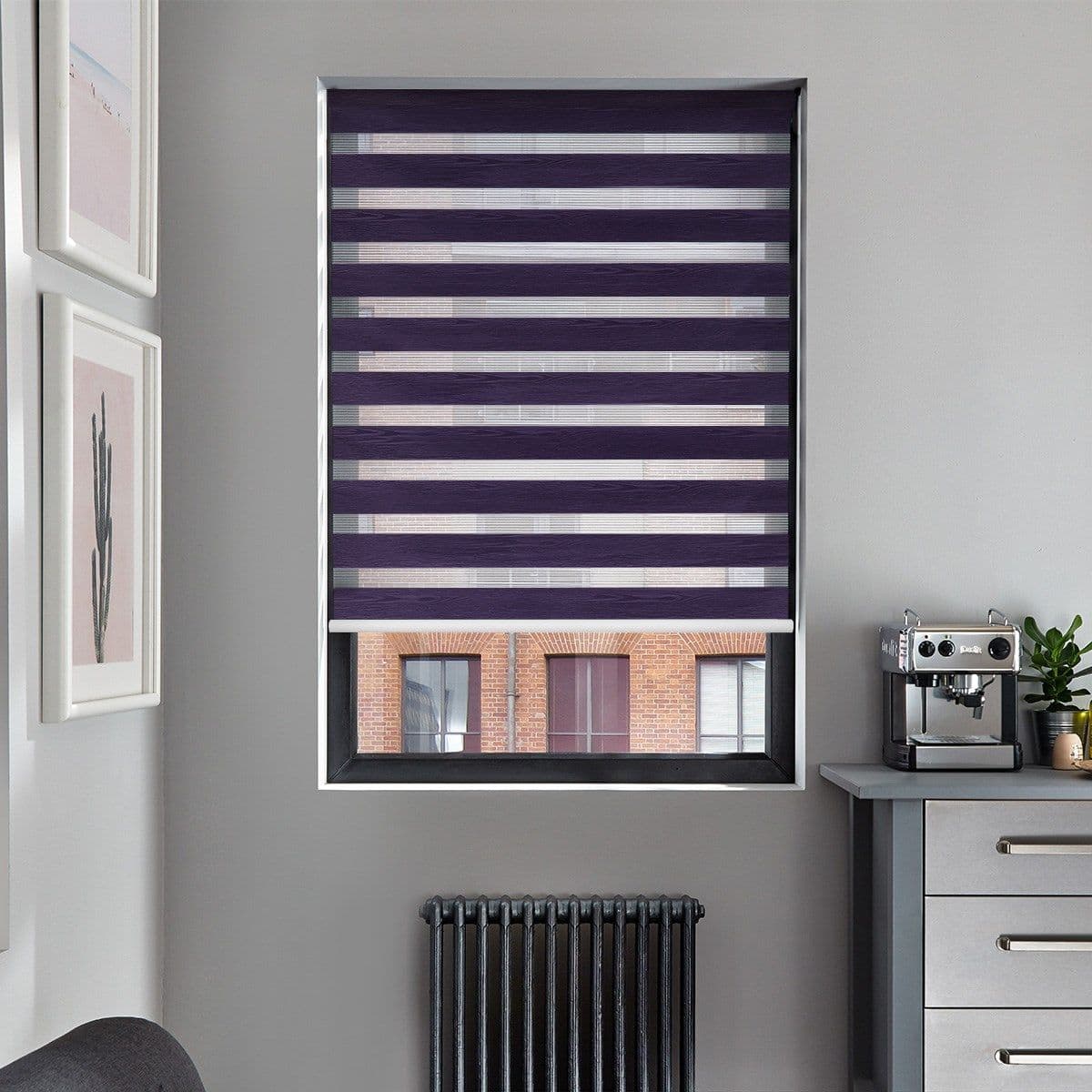 Zebra roller blind, purple and sheer alternating horizontal stripes, lowered to cover most of the window and partially filter light; modern gray kitchen wall with coffee machine, radiator, framed prints.