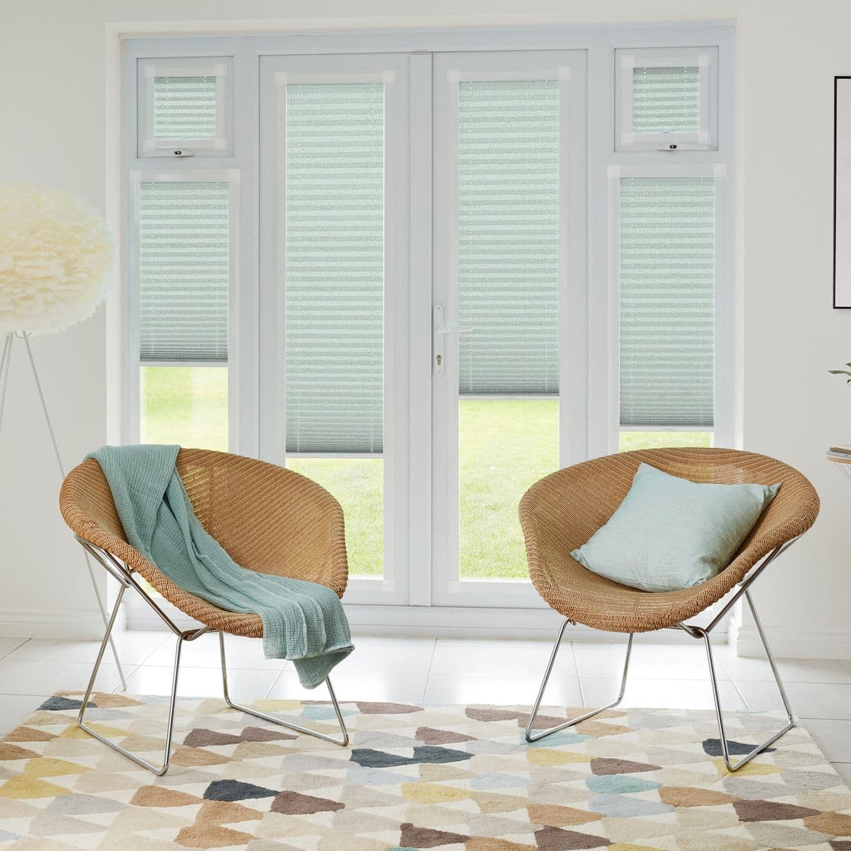 Pleated cellular blinds (seafoam with horizontal texture) covering French doors and sidelights, lowered midâ€‘height and filtering daylight; modern bright sitting area with two rattan chairs, patterned rug, lawn view.