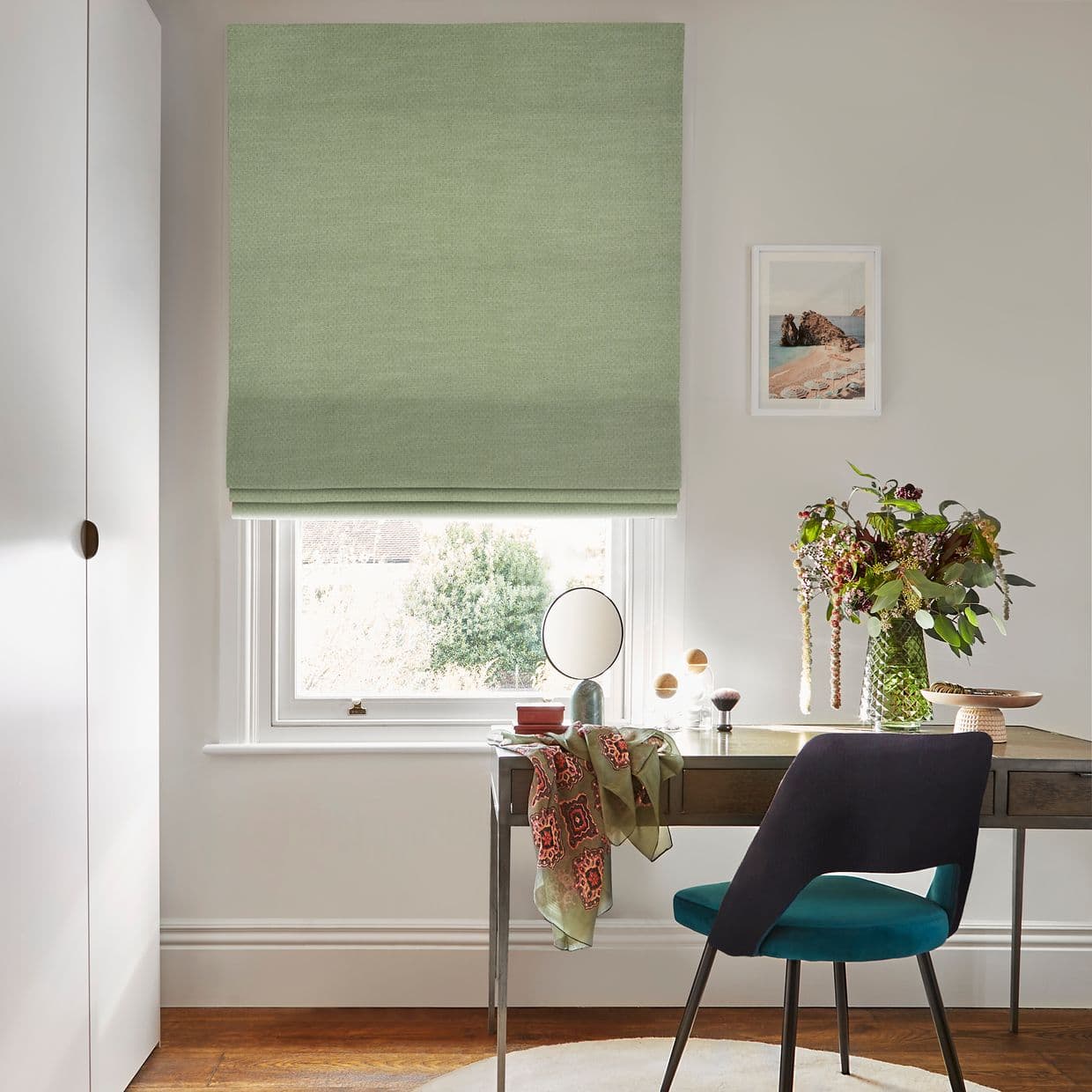 Pale green Roman blind covering the window, partly lowered and filtering daylight; small desk beneath with mirror, teal chair and vase of flowers in a softly lit home office.