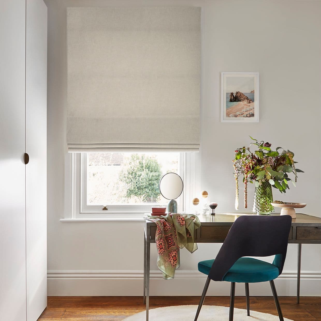 Beige fabric Roman blind, lowered with folds and filtering daylight, covering a window; in a bright neutral home office with a wooden desk, mirror, vase of foliage and teal chair.