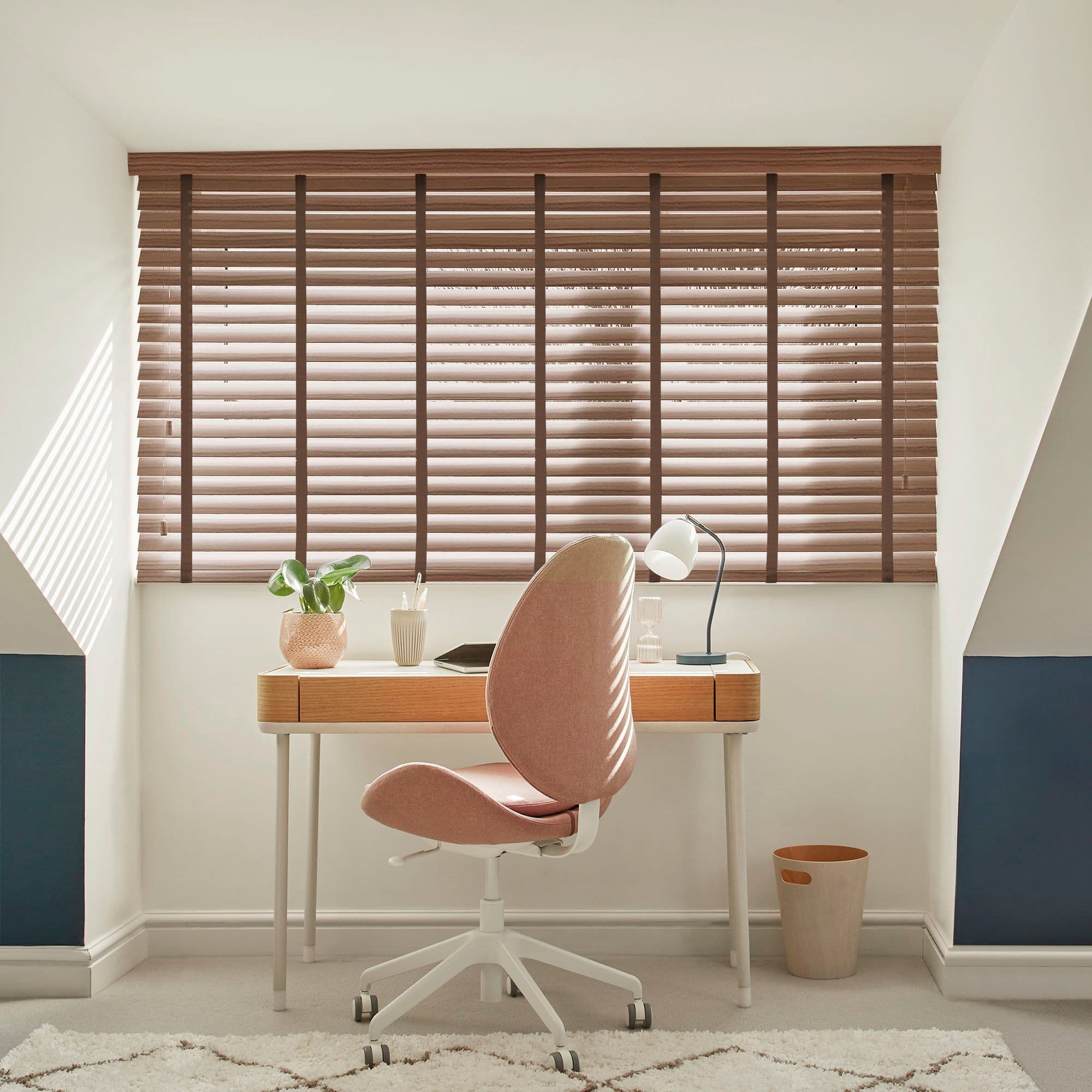 Wooden horizontal blinds covering a wide window, brown slats slightly tilted to filter sunlight; a small wooden desk with plant, lamp and a pink swivel chair in a bright attic-style home office.