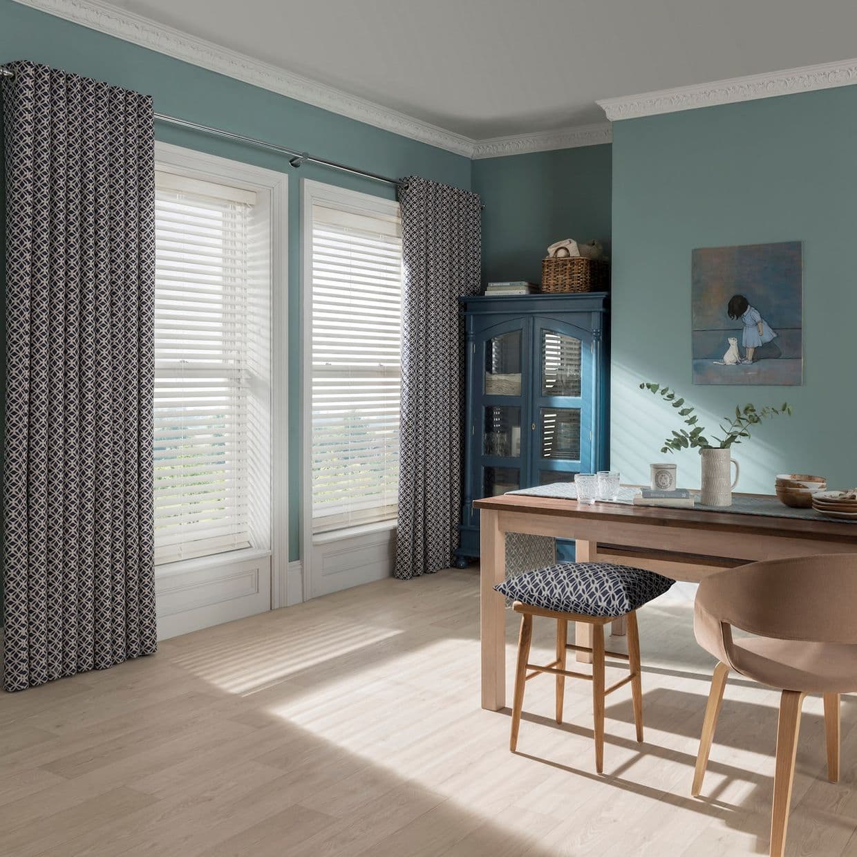 Patterned curtains and white horizontal blinds at tall windows, blinds filtering sunlight through slats â€” casting striped light across pale wood floor in teal-walled dining room with table and blue cabinet.