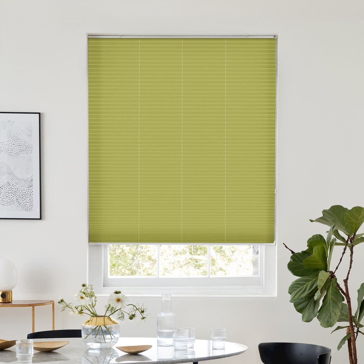 Green pleated cellular blind, fully lowered across the window, filtering light through a narrow bottom gap; above a dining table with glassware, framed art, and a potted fiddleâ€‘leaf fig.