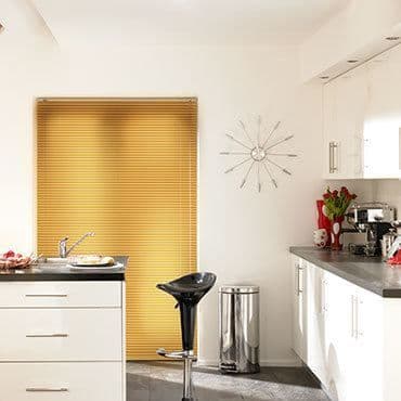 Yellow pleated blinds mounted in the window, fully closed and softly filtering sunlight; modern white kitchen with island, black barstool, stainless trash can, countertop appliances and red tulips.