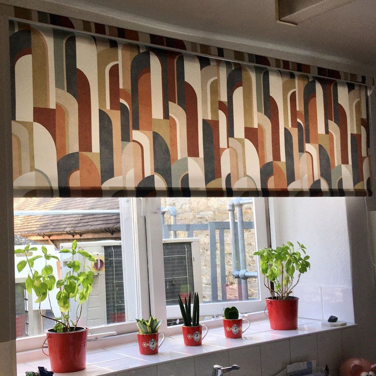 Retro-arched patterned roller blind, lowered across the window, filtering light; kitchen windowsill beneath holds red potted herbs and small succulents, outside view of a stone wall and metal pipes.