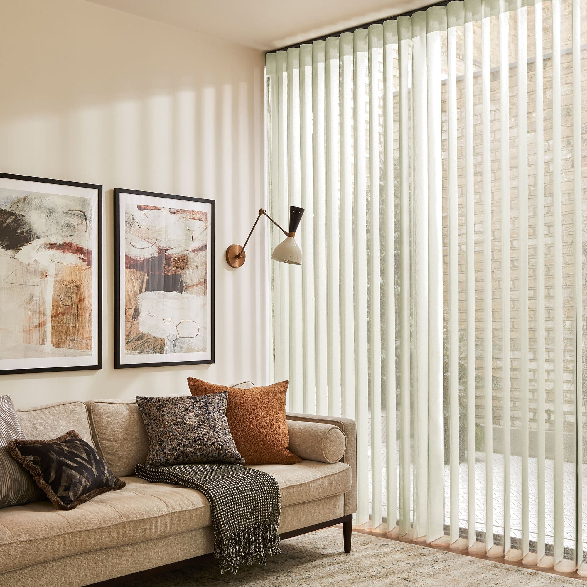 Sheer cream vertical blinds covering a full-height window, filtering sunlight into soft striped shadows across a living room with a beige sofa, textured cushions and framed wall art.