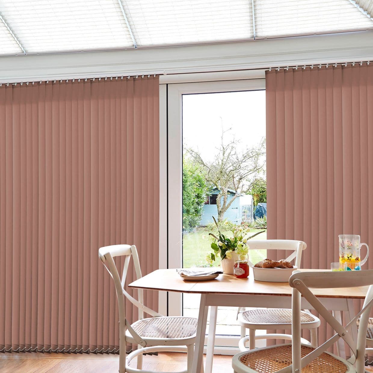 Vertical rose-beige vertical blinds covering a patio sliding glass door, mostly closed with a central gap; filtering daylight into a sunlit dining area with table, chairs, flowers and garden view.