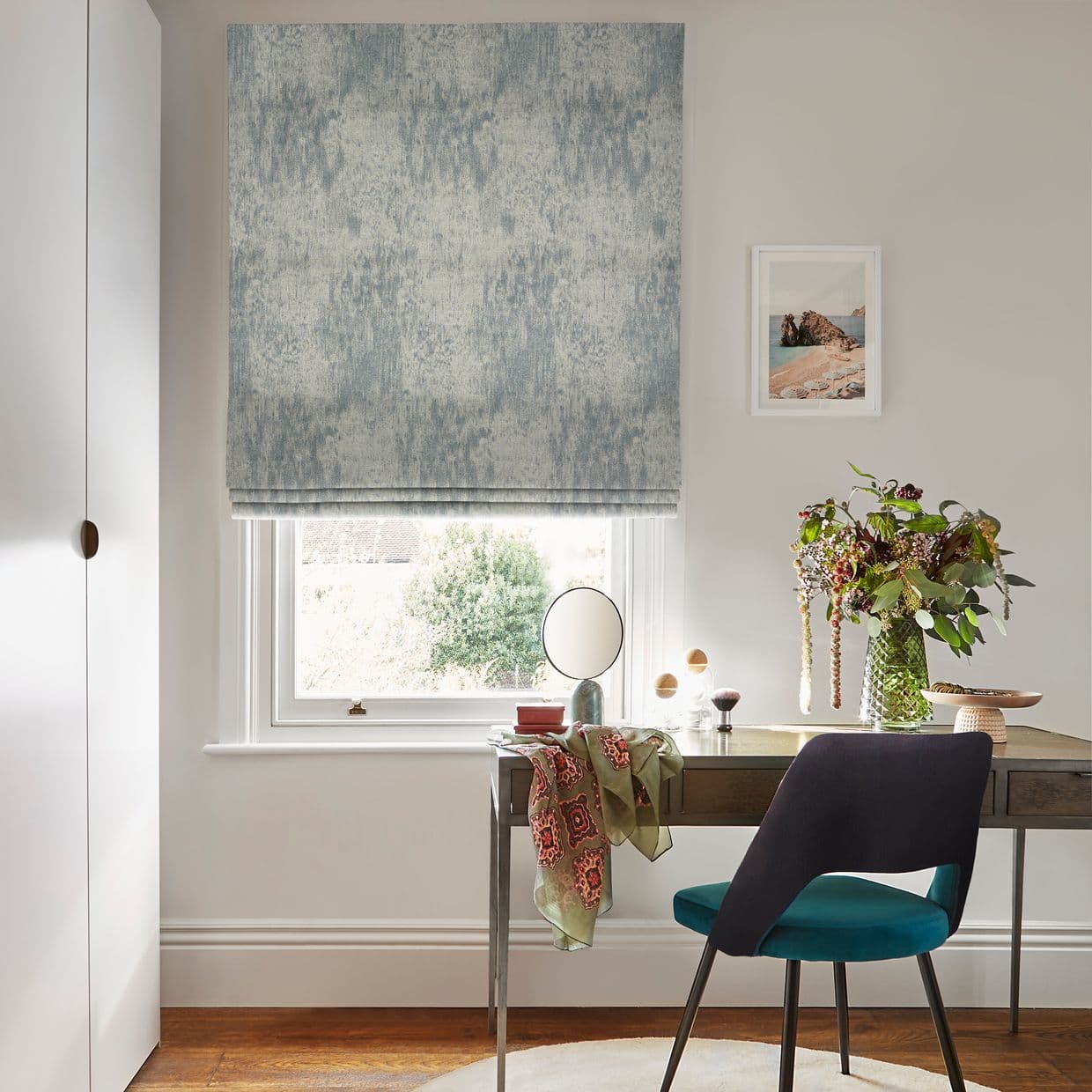 A textured blue-gray Roman blind hangs fully lowered, softly filtering daylight over a desk; room includes a window-facing wooden desk with vase of flowers, teal chair, and framed seaside print.