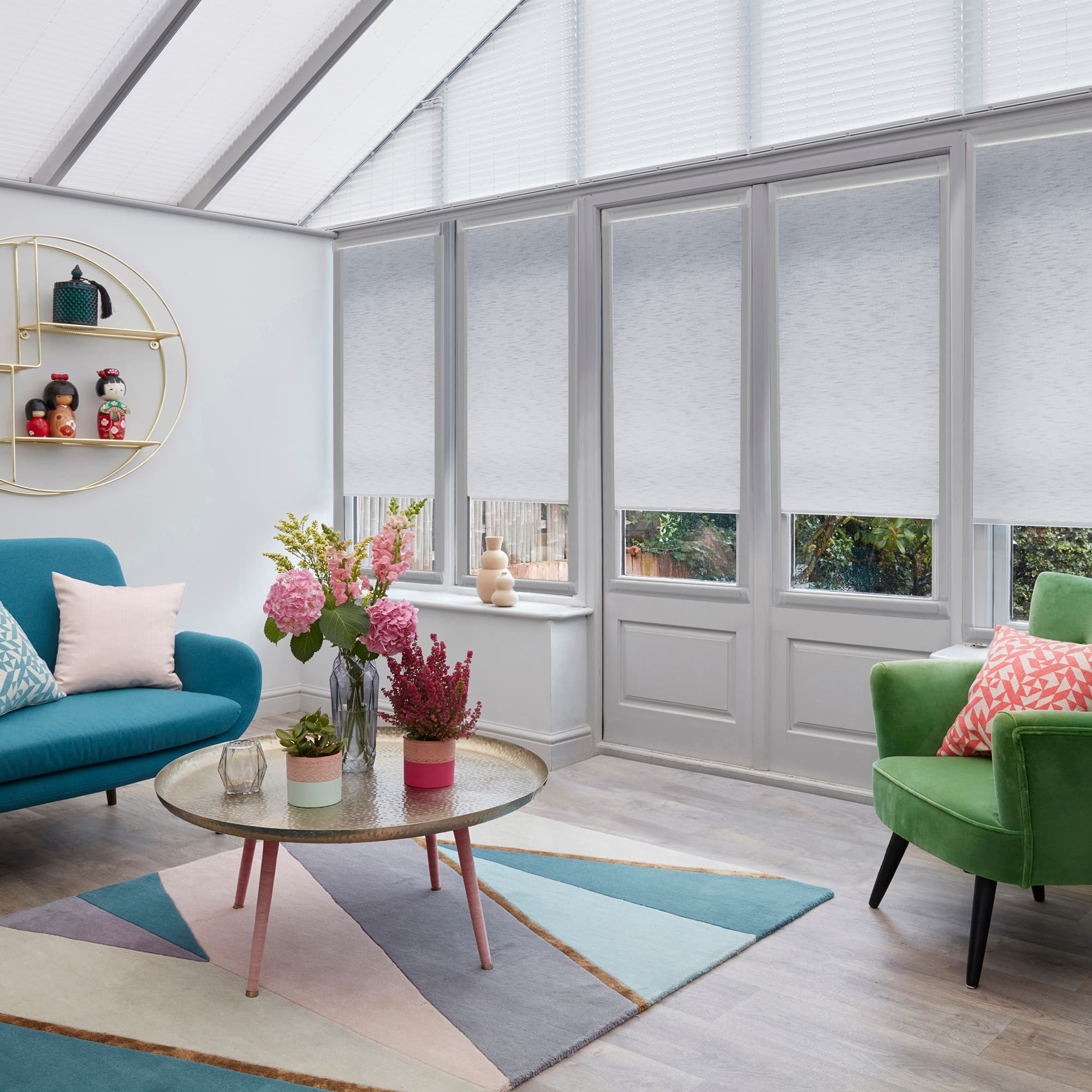 White roller blinds and pleated roof blinds covering tall glass doors and windows, partially lowered and filtering soft daylight in a bright conservatory with colorful seating, geometric rug and plants.