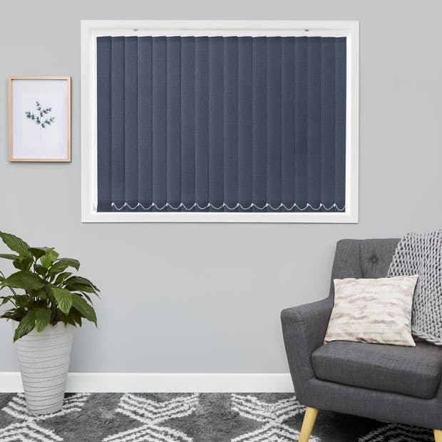 Navy vertical blinds covering a white framed window, closed and light-blocking with a scalloped stitched hem; in a gray living room with a dark armchair, potted plant, framed art, and patterned rug.