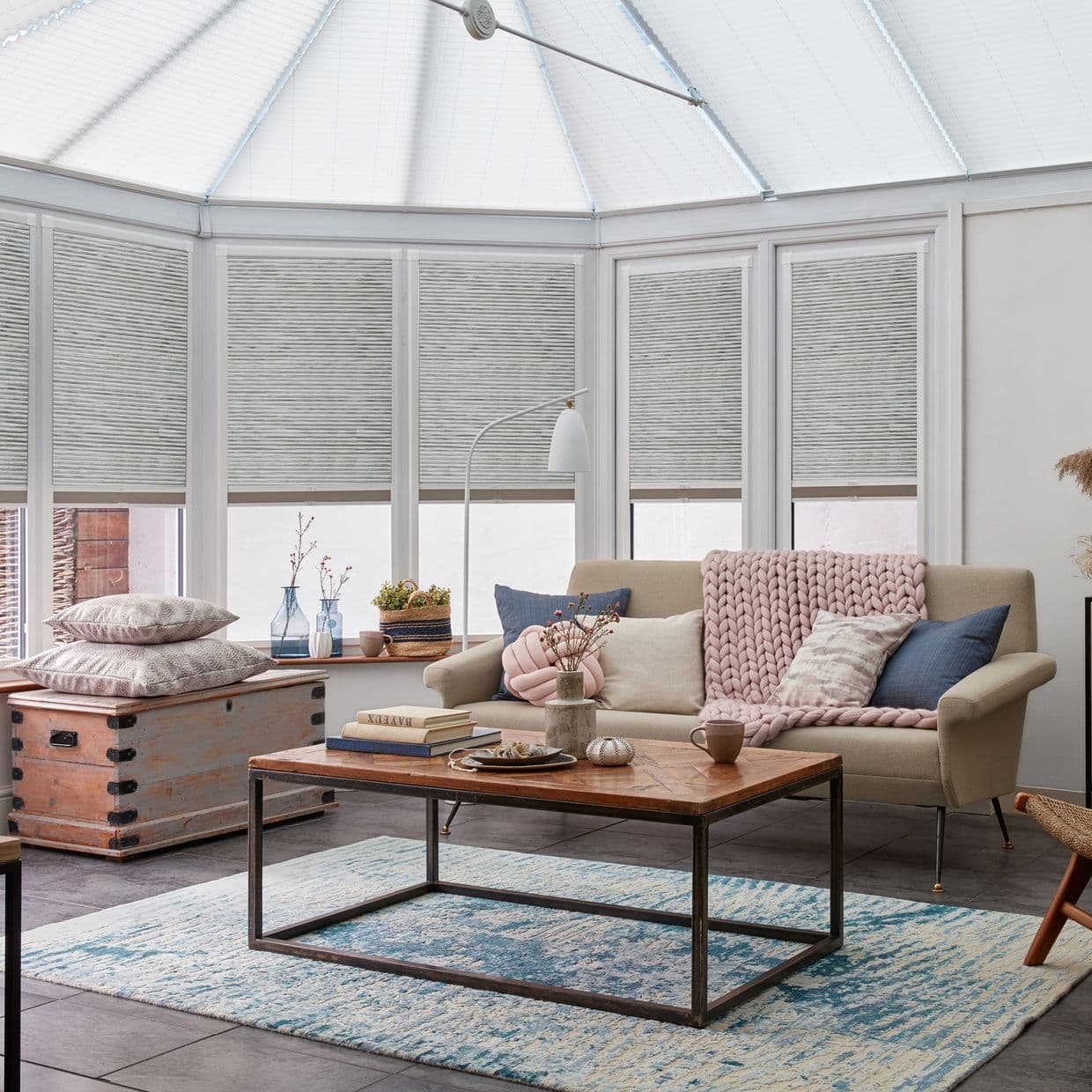 Gray pleated cellular blinds, lowered across bay conservatory windows with a small clear strip at the bottom, filtering soft daylight; cozy sunroom with a sofa, wooden coffee table, and pale decor.