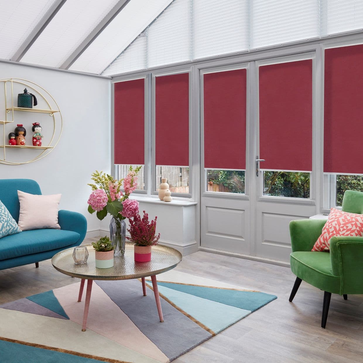 Maroon roller blinds on tall French windows and door, lowered over lower panels while white pleated blinds shade the slanted roof, blocking light and view; bright conservatory with colorful seating.