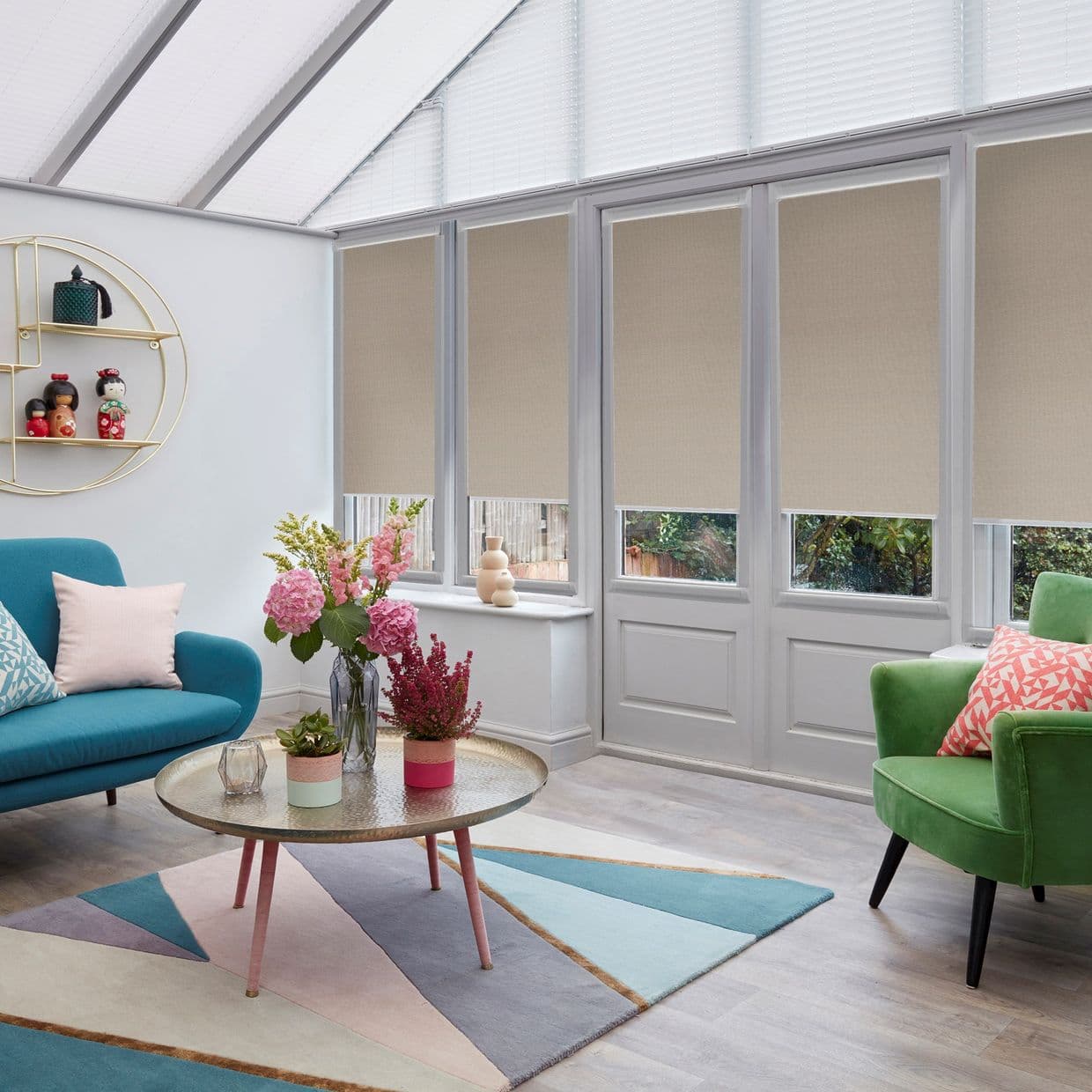 Beige roller blinds lowered across tall glazed doors and sloping roof windows, softly filtering daylight; in a bright conservatory-style living room with teal and green chairs, coffee table, and flowers.
